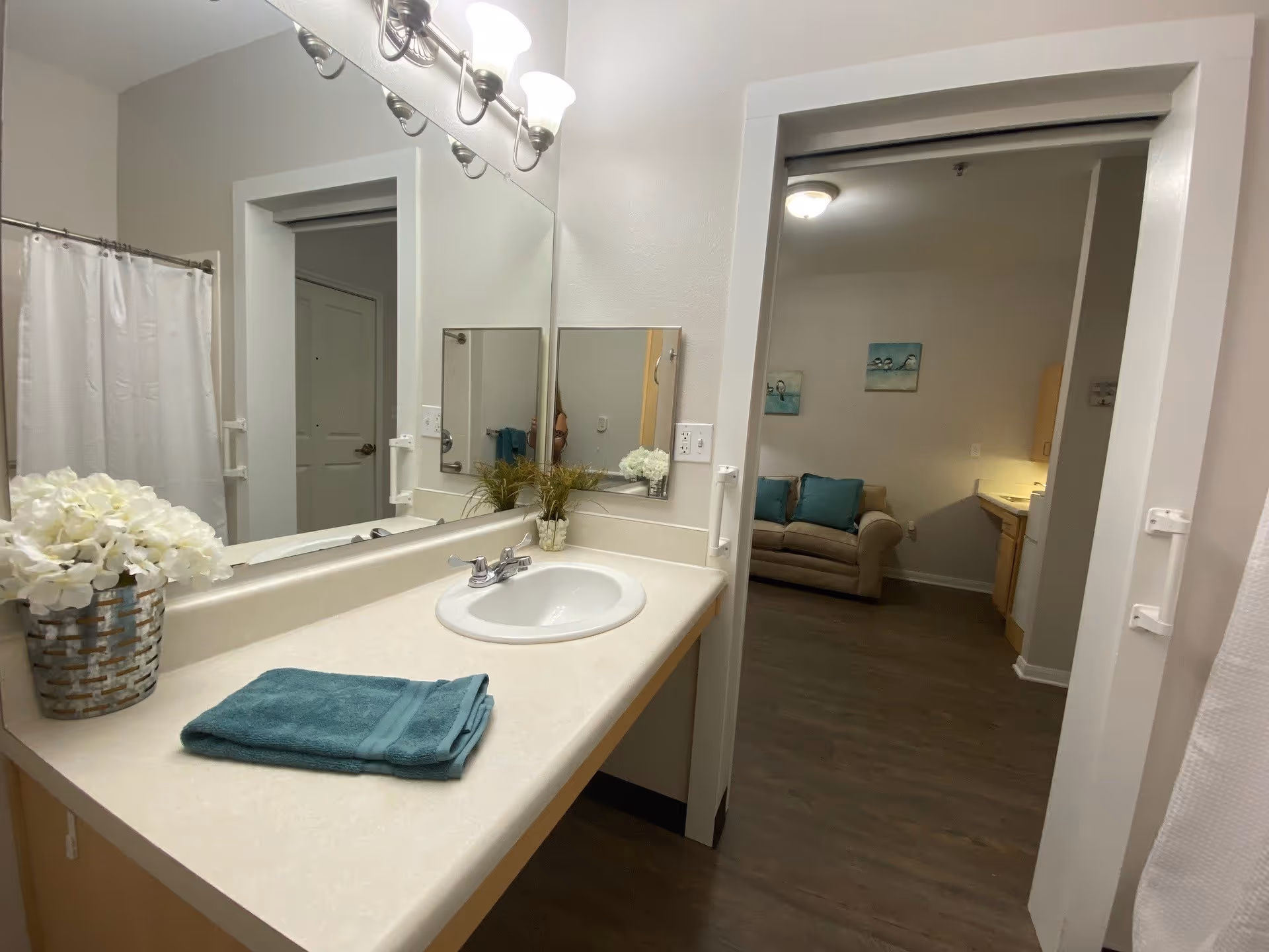 A bathroom vanity with a sink, a folded teal towel, and a vase of white flowers on the countertop. A large mirror with lights above it reflects the bathroom and an open doorway leading to a living area with a beige couch and teal pillows.