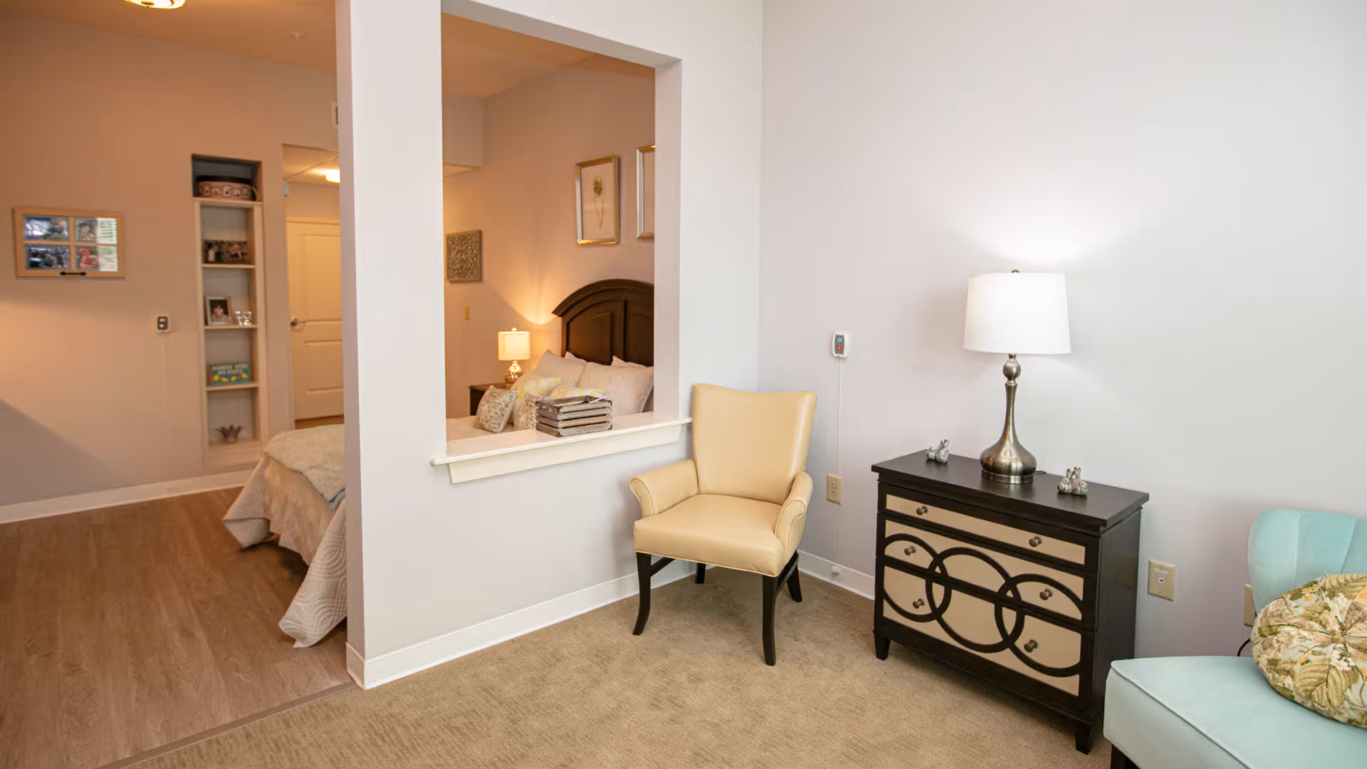 Interior view of a senior living facility showing a cozy sitting area with a beige armchair, a black and beige dresser with a lamp on top, and a light blue chair with a floral cushion. Through a large open window in the wall, a bedroom with a bed, nightstand, and wall art is visible.