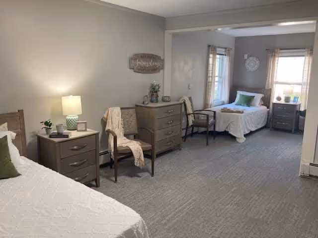 A spacious bedroom with two single beds, each with white bedding and green pillows. Between the beds are wooden nightstands with lamps and decorative items. There are two chairs with light-colored throws draped over them, and a wooden dresser with plants and decor. The room has light gray walls, carpeted floor, and windows with curtains allowing natural light.