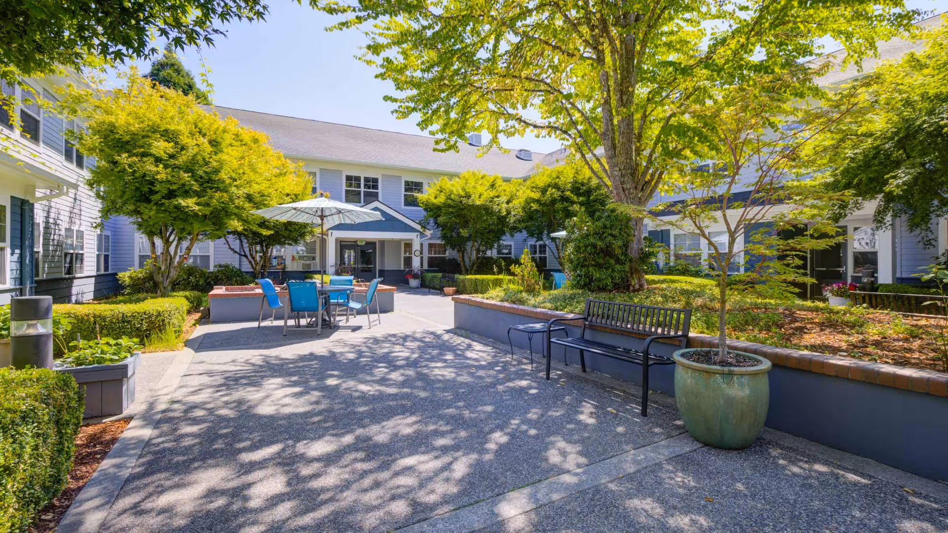 Sunny landscaped courtyard with outdoor tables and chairs under an umbrella, benches, potted plants, and a surrounding two-story building.