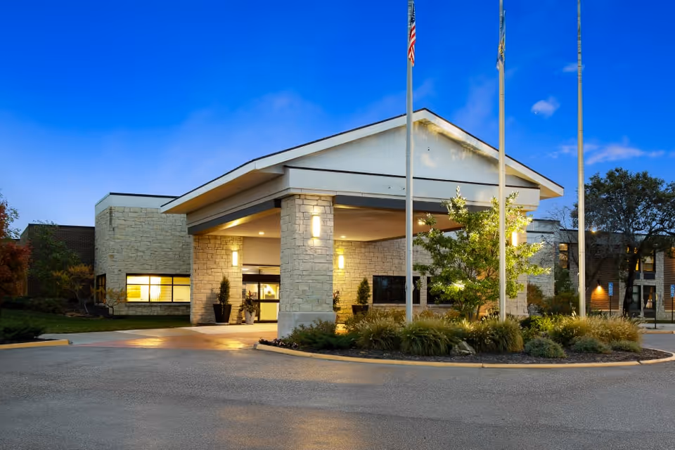 Front entrance of a senior living building with a covered porte-cochere, stone facade, landscaping, and flagpoles at dusk.