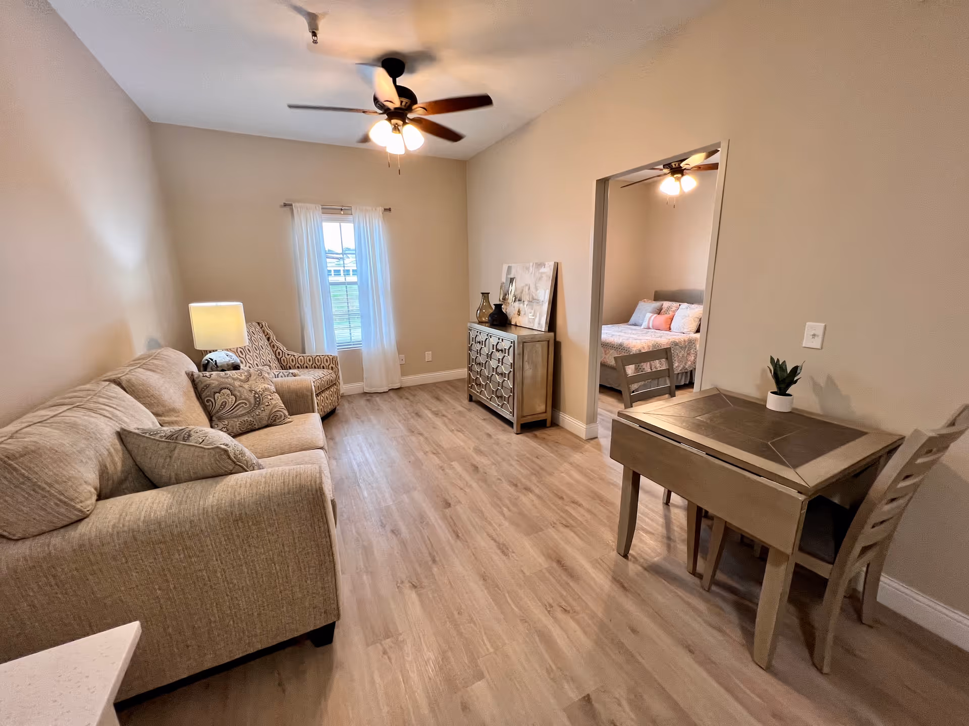 A cozy living room with a beige sofa and patterned armchair near a window with white curtains. A wooden sideboard with decorative items is against the wall. A small dining table with two chairs and a potted plant is on the right. Through an open doorway, a bedroom with a bed and ceiling fan is visible. The room has light wood flooring and neutral-colored walls.