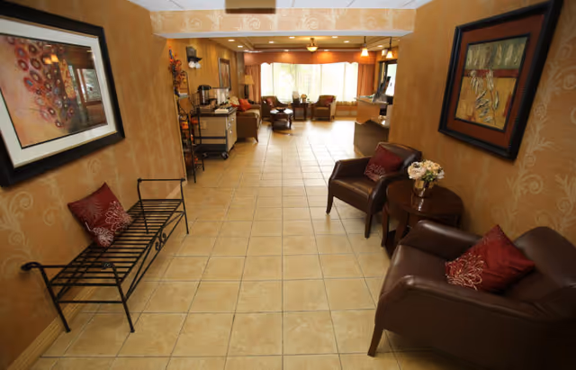 A spacious interior room with tiled flooring and warm beige walls decorated with framed artwork. The room features two brown leather armchairs with red decorative pillows and a small round wooden table with a flower arrangement between them. On the left side, there is a black metal bench with a red pillow. In the background, there is a seating area with multiple chairs and a large window letting in natural light.