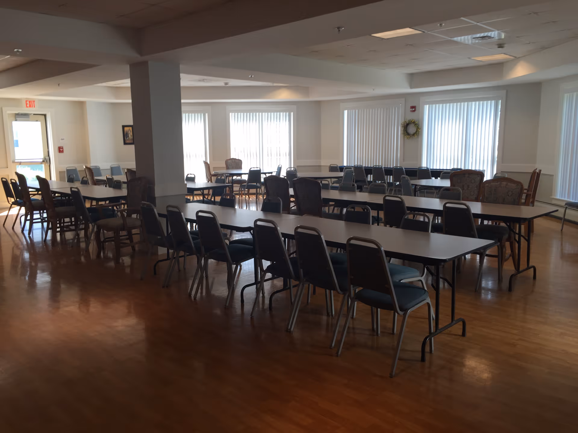 A spacious dining room with multiple long tables and chairs arranged neatly. The room has large windows with vertical blinds allowing natural light to enter. The floor is wooden, and there is a white column in the middle of the room. An exit door is visible on the left side.