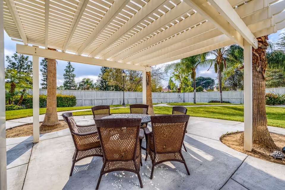 Outdoor patio area with a white pergola overhead, featuring a round table surrounded by six wicker chairs. The patio is set on a concrete surface with green grass, palm trees, and other plants visible in the background, enclosed by a white fence.