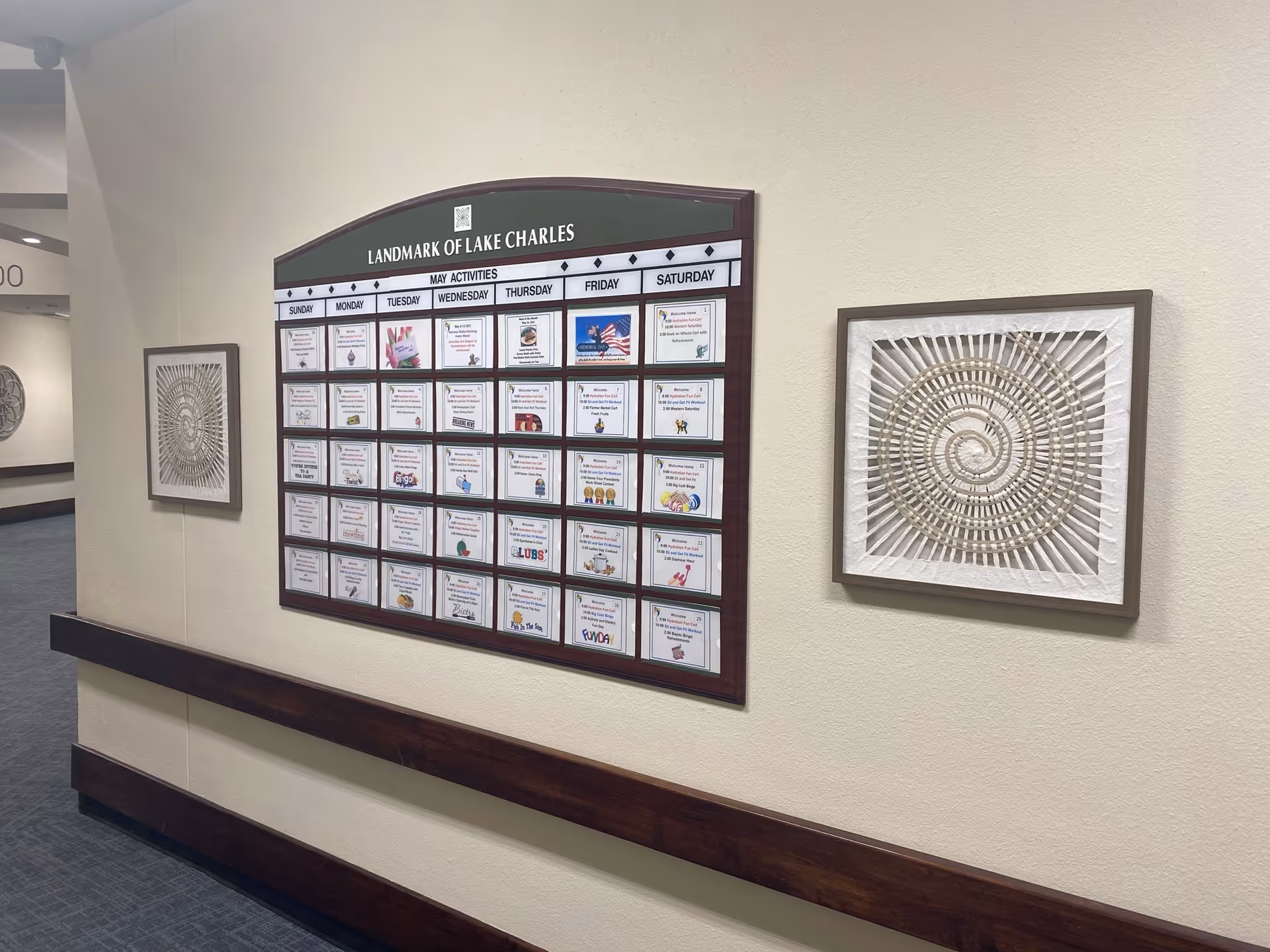 A hallway wall inside Landmark of Lake Charles featuring a large monthly activities calendar for May, flanked by two framed decorative art pieces with spiral designs. The wall is painted beige and has a wooden handrail along it.