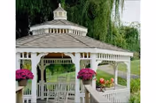 White octagonal gazebo with rocking chairs and potted flowers on a green lawn with trees in the background.