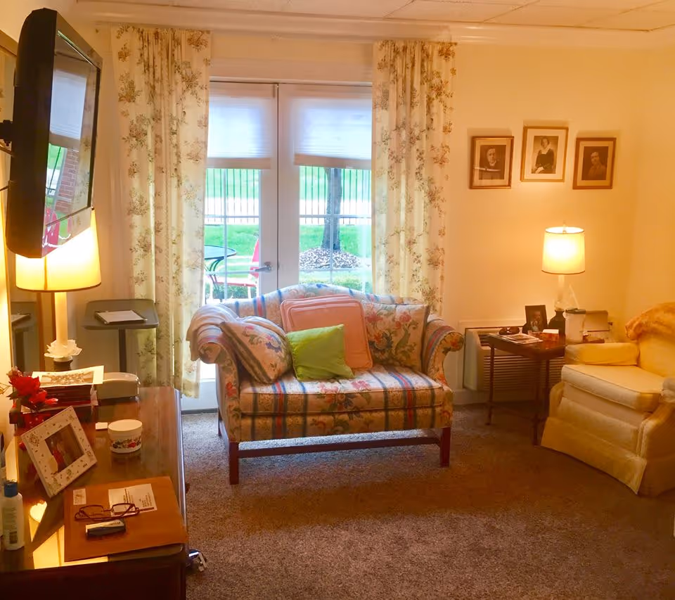 A cozy living room in Mari De Villa Nursing Home featuring a floral-patterned loveseat with colorful cushions, a yellow armchair, a wooden side table with a lamp, framed pictures on the wall, and a TV mounted on the wall. French doors with floral curtains open to a green outdoor area.