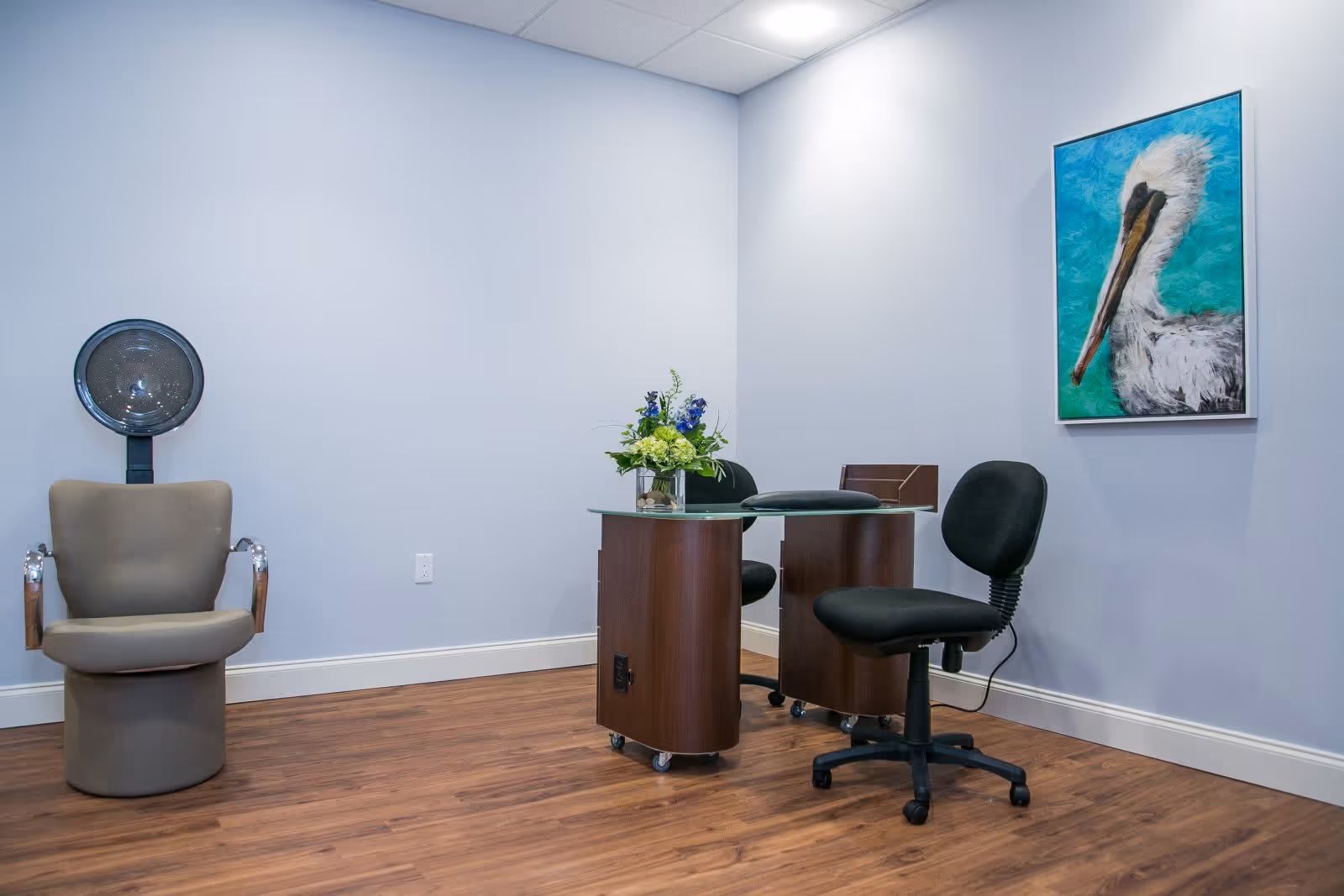 A small room with light blue walls and wood flooring featuring a salon hair dryer chair on the left, a small glass-top desk with two black office chairs on wheels, a flower arrangement on the desk, and a framed painting of a pelican on the wall.