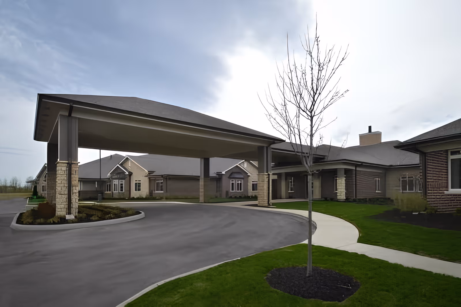 Exterior view of The Ganzhorn Suites showing a covered driveway entrance with stone pillars, a curved sidewalk, a small tree with no leaves, and well-maintained green grass surrounding the building under a cloudy sky.