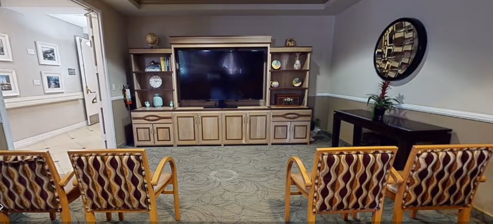 A common area with four patterned chairs facing a large flat-screen TV mounted on a wooden entertainment center with shelves holding decorative items. To the right, there is a dark console table with a plant and a round decorative mirror above it. The walls are painted beige with white trim, and there is an open doorway leading to a hallway.