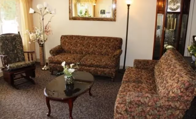 A cozy living room with two floral patterned sofas, a matching armchair, a wooden coffee table with a small flower arrangement, a tall floor lamp, a large wall mirror, and a grandfather clock in the corner.