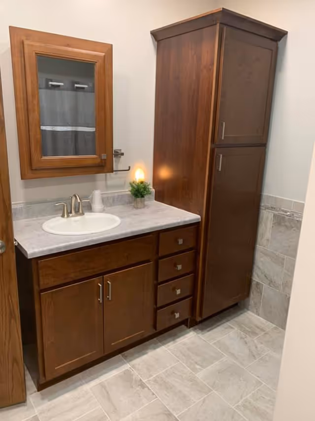 Bathroom vanity with wood cabinets, a marble countertop and sink, wall mirror, tall storage cabinet, and tiled floor.