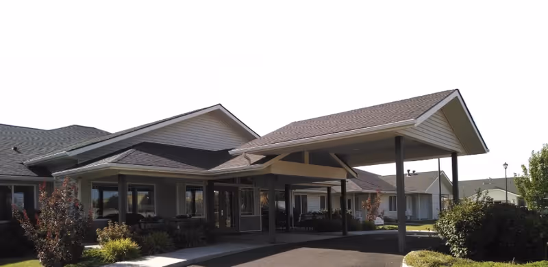 Front entrance of a single-story senior living building with a covered porte-cochère, columns, and surrounding shrubs.