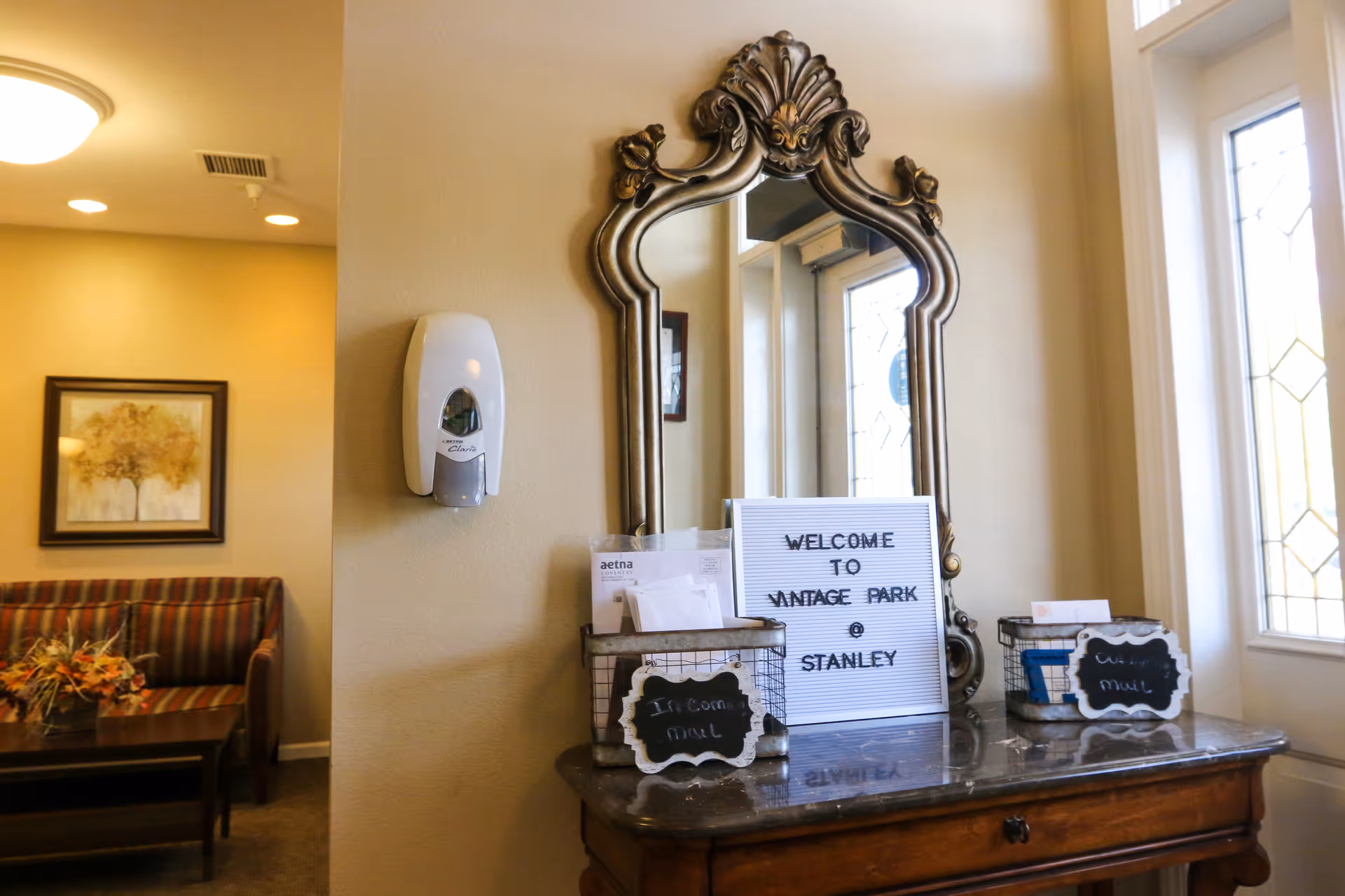 Ornate mirror over an entry table with a welcome sign and mail baskets in a senior living facility lobby.