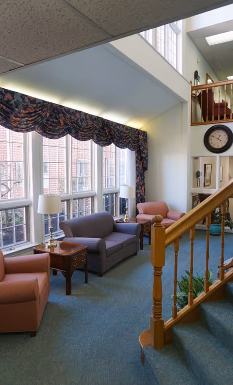 A cozy sitting area with large windows covered by floral valances, featuring a purple loveseat, two pink armchairs, wooden side tables with lamps, a staircase with wooden railing, and a large wall clock above a small hallway.