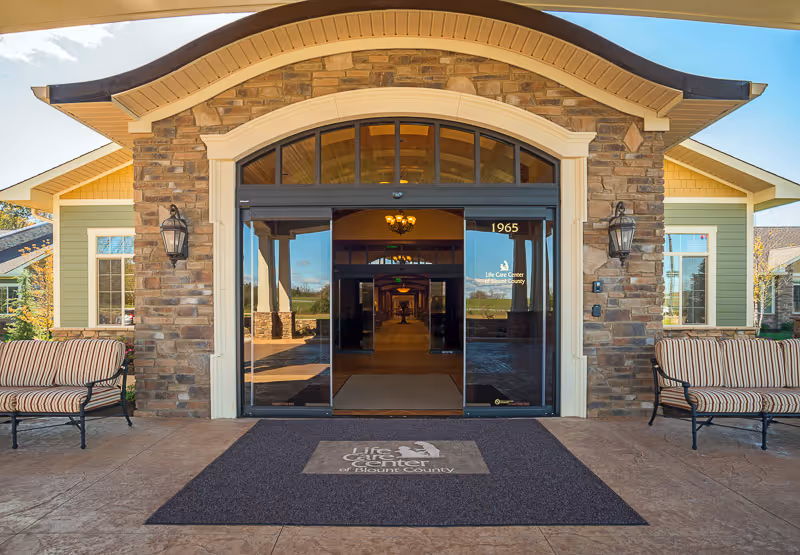 Entrance of Life Care Center of Blount County featuring a stone and wood exterior with large glass doors. Two striped cushioned benches are placed on either side of the entrance. A dark mat with the facility's logo is laid out in front of the doors, and the building number 1965 is visible above the door.