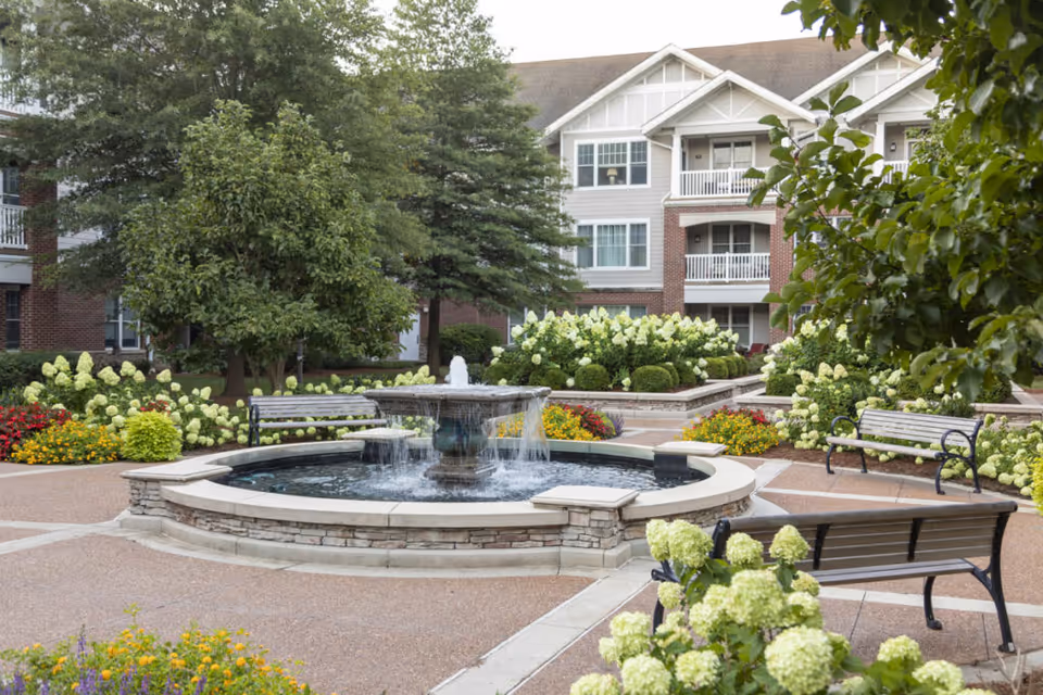 Outdoor courtyard area with a central stone fountain surrounded by benches, colorful flower beds, and green trees, with a multi-story residential building in the background.