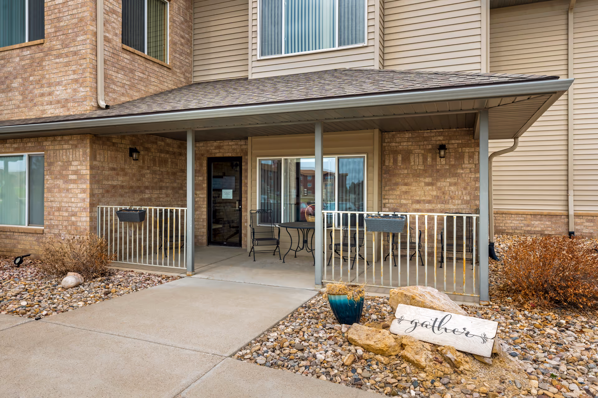 Covered patio area outside a senior living facility with a small table and chairs, beige brick and siding exterior walls, a concrete walkway, decorative rocks, a blue planter, and a wooden sign that says 'gather'.