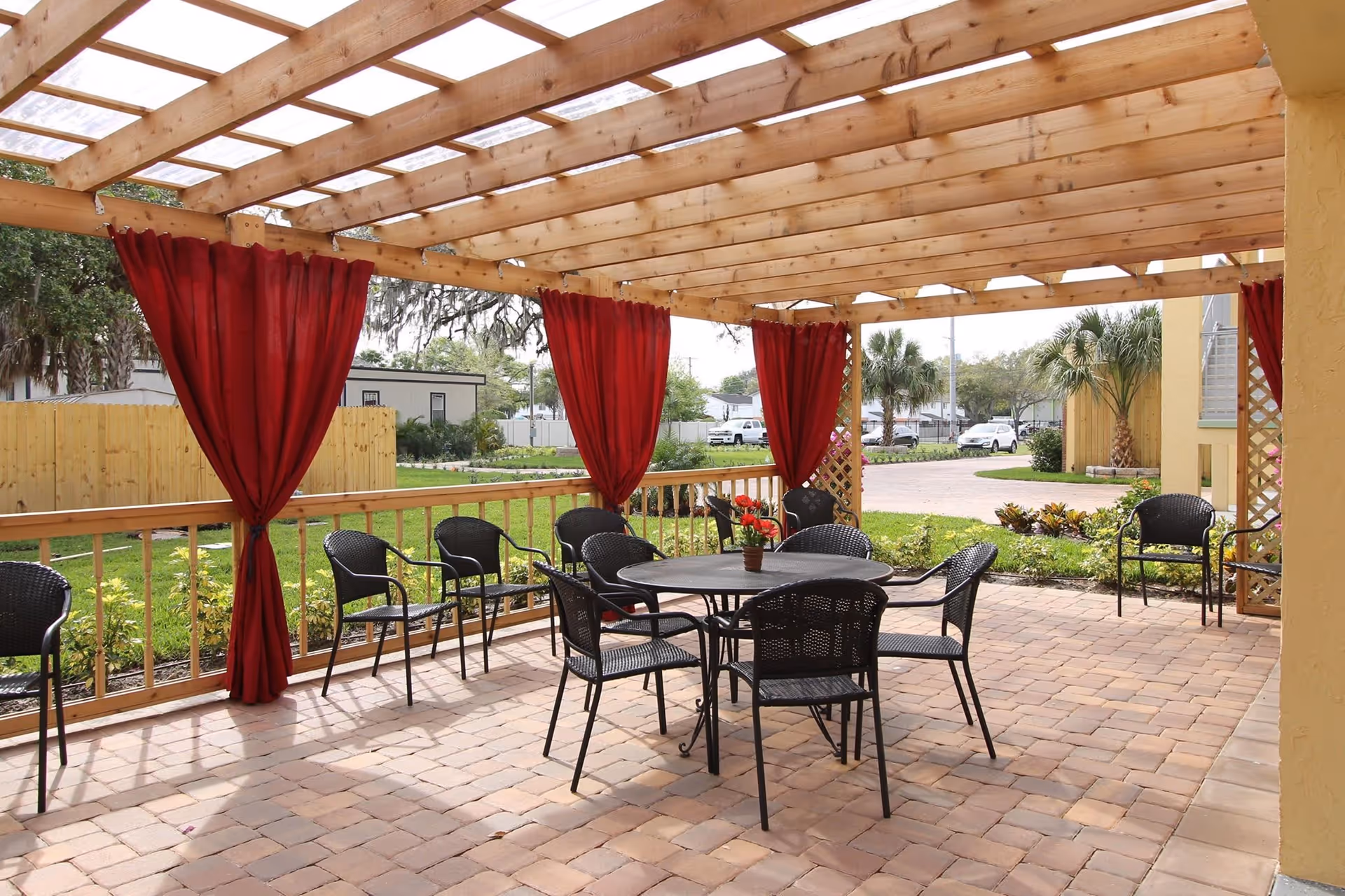 Outdoor patio area with a wooden pergola overhead, red curtains tied back to wooden posts, a round table with a small potted plant in the center, and several black metal chairs arranged around the table and along the railing. The patio floor is made of brick pavers, and there is greenery and a parking area visible in the background.