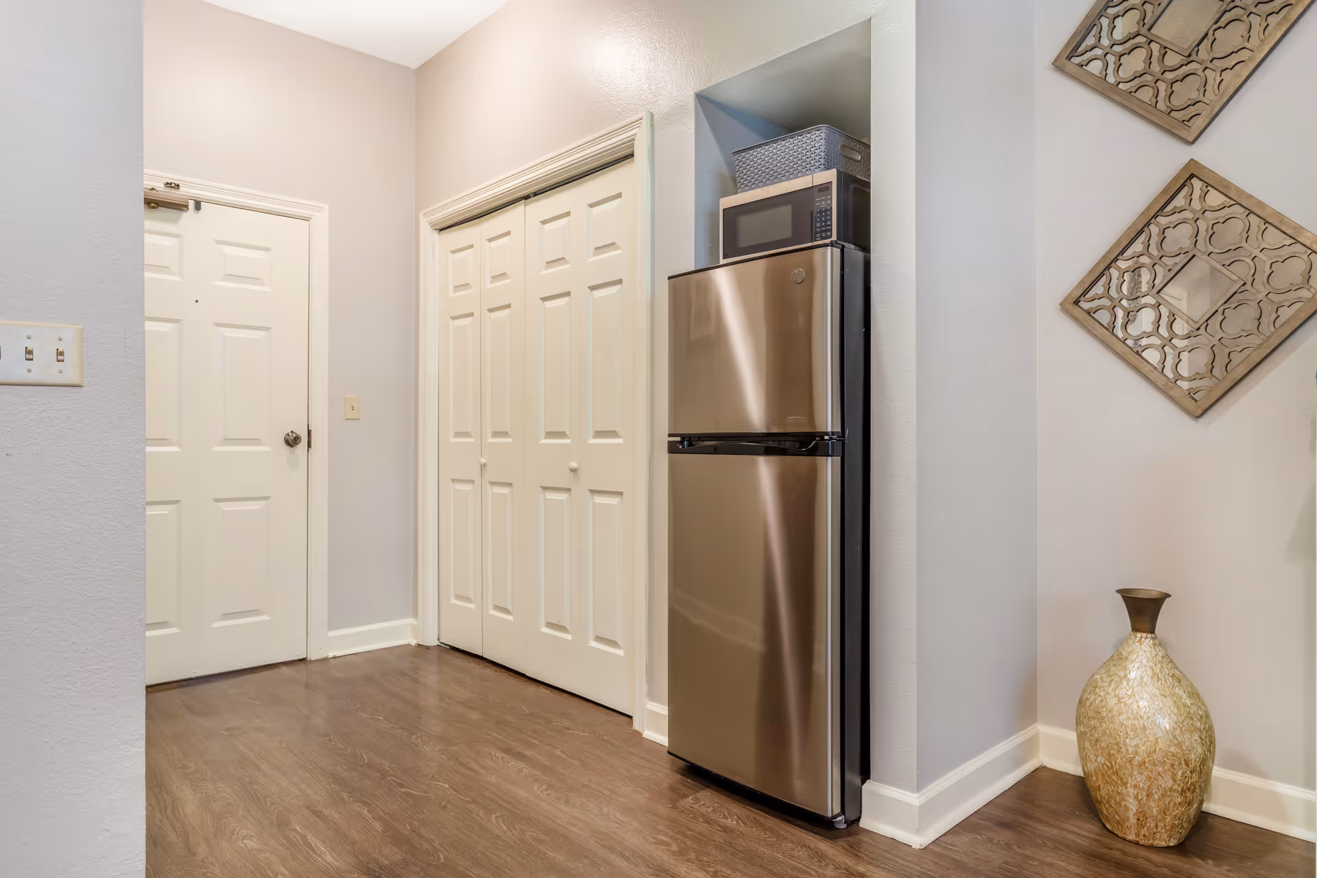 Entry hallway with a stainless steel refrigerator and microwave beside double closet doors, decorative wall art, and a floor vase.