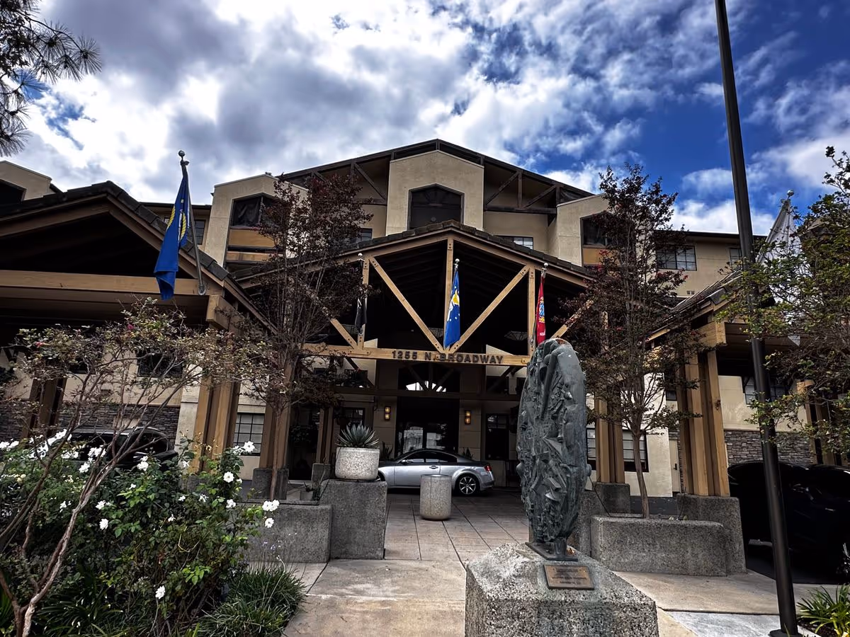 Front entrance of Cypress Court Senior Living showing a covered porte-cochere with flags, a sculpture in the foreground, planters and a car under the canopy.