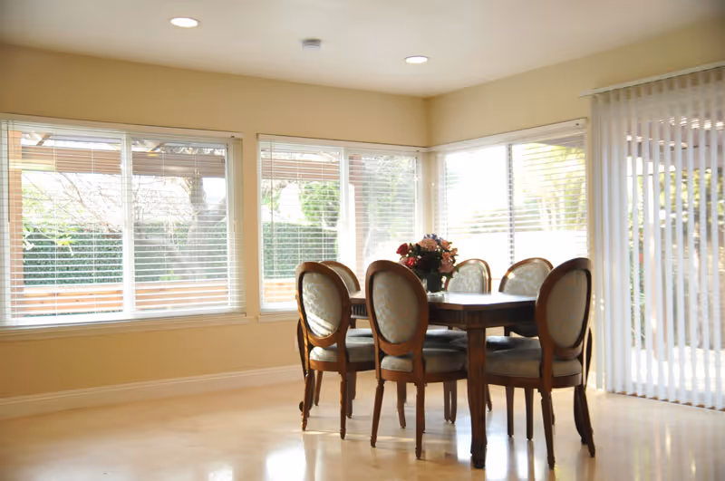 A bright dining area with a wooden dining table and six upholstered chairs. The room has large windows with white blinds and vertical blinds on a sliding glass door, allowing natural light to fill the space. A floral centerpiece is placed on the table.