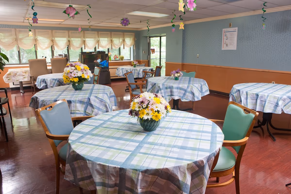 A dining room with several round tables covered in blue and green plaid tablecloths. Each table has a vase with a colorful bouquet of flowers. The room has large windows with sheer curtains, wooden chairs with green and blue cushions, and decorations hanging from the ceiling. The floor is a polished reddish-brown wood.