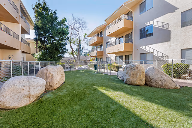 Sunlit courtyard between multi-story apartment buildings with balconies, a fenced artificial turf lawn and several large boulders.