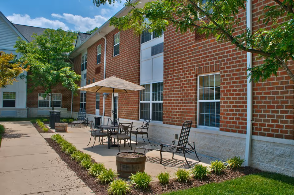 Outdoor patio area at Arbor Crest of Silver Spring featuring a brick building with several windows, a table with an umbrella, multiple chairs, a lounge chair, and landscaped greenery along a paved walkway under a clear blue sky.