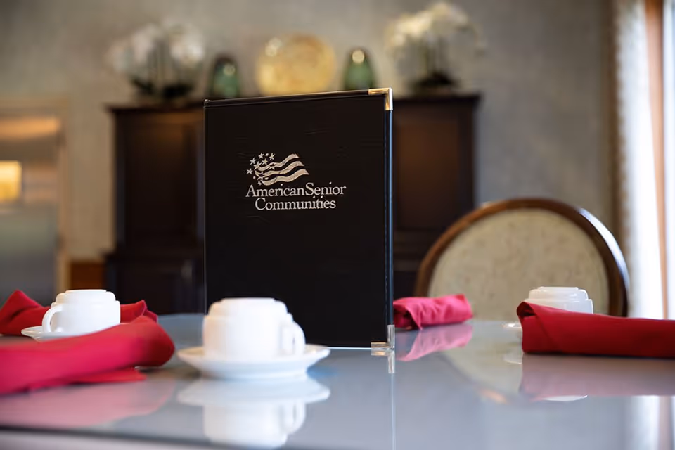 A dining table set with white cups and saucers, red cloth napkins, and a black menu holder with the logo and text 'American Senior Communities' in the center. The background shows a blurred cabinet and a cushioned chair.