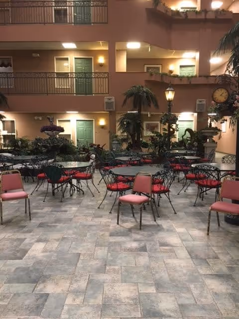 Indoor seating area with multiple round tables and chairs with red cushions, surrounded by plants and decorative elements. The background shows multiple floors with green doors and railings, resembling an atrium or lobby area of a senior living facility.