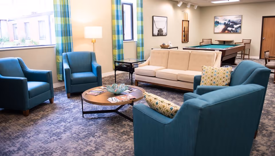 A bright and spacious living room area with a beige sofa, three blue armchairs, a round wooden coffee table with magazines, and a floor lamp near the window. The room features large windows with blue and green plaid curtains, a pool table in the background, and framed artwork on the walls.