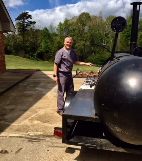 A man wearing gray scrubs stands outdoors next to a large black barbecue smoker on a concrete driveway with green trees and grass in the background under a partly cloudy sky.
