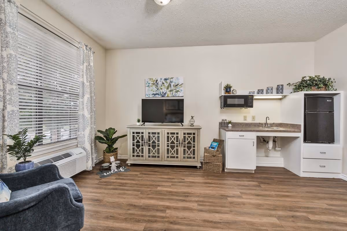 A cozy senior living room with a blue armchair near a window with patterned curtains. There is a white cabinet with a flat-screen TV on top, a small indoor plant, and a decorative piece. To the right, there is a kitchenette area with a microwave, sink, countertop, and a black mini refrigerator. The floor is wood-style laminate, and the walls are painted white.