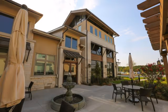 Outdoor patio area of a senior living facility with a stone and beige exterior building, several windows, a small water fountain, patio tables with chairs, and closed umbrellas under a clear sky.