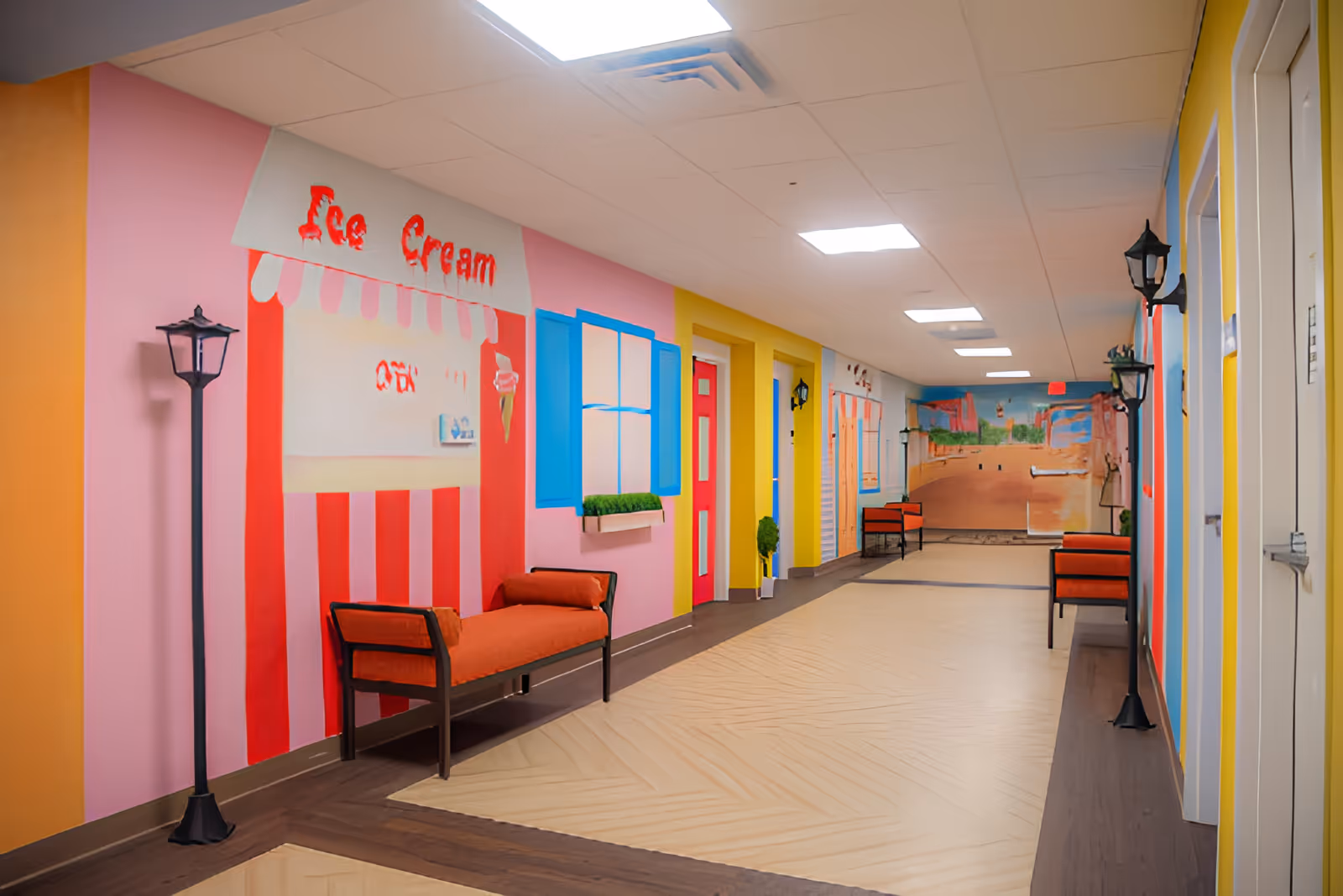A brightly colored indoor hallway with walls painted to resemble storefronts, including an ice cream shop with red and white stripes and a blue window with a flower box. The hallway has benches with orange cushions, black lamp posts mounted on the walls, and a mural at the far end depicting an outdoor scene.