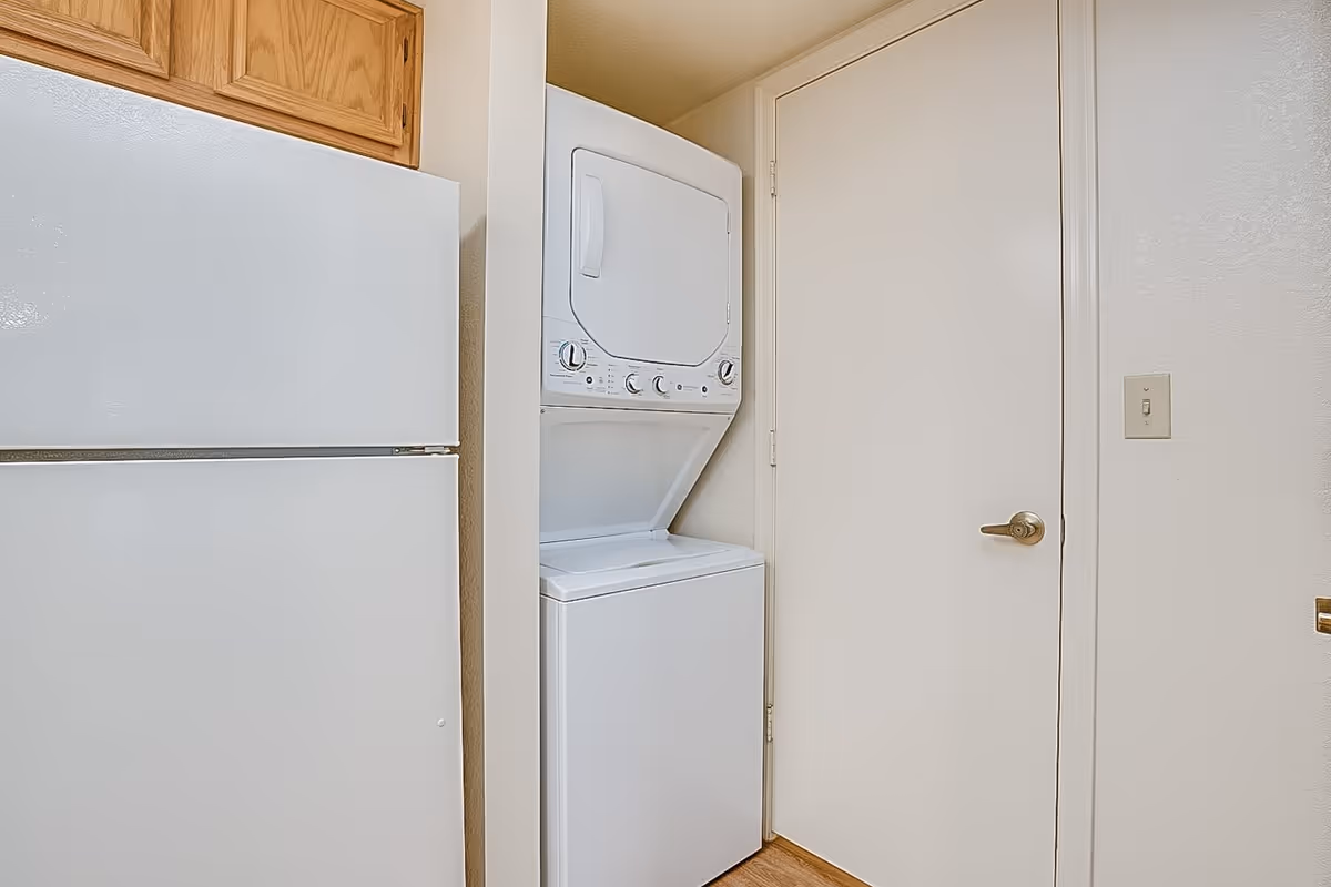 A compact laundry area featuring a stacked white washer and dryer unit next to a white refrigerator with wooden cabinets above, set against beige walls and a closed white door with a silver handle.