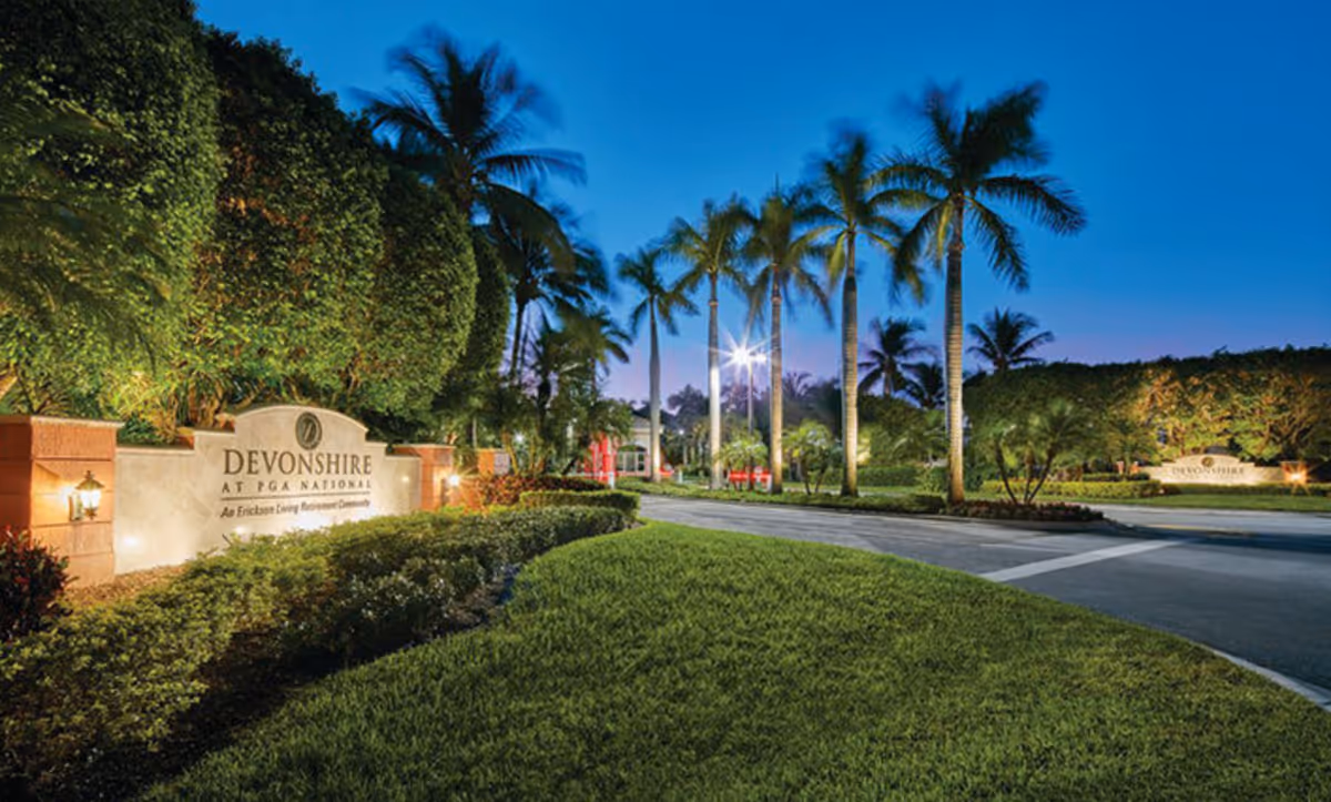 Entrance to Devonshire Senior Living Community at PGA National during twilight, featuring a well-lit sign, manicured bushes, palm trees, and a paved driveway.