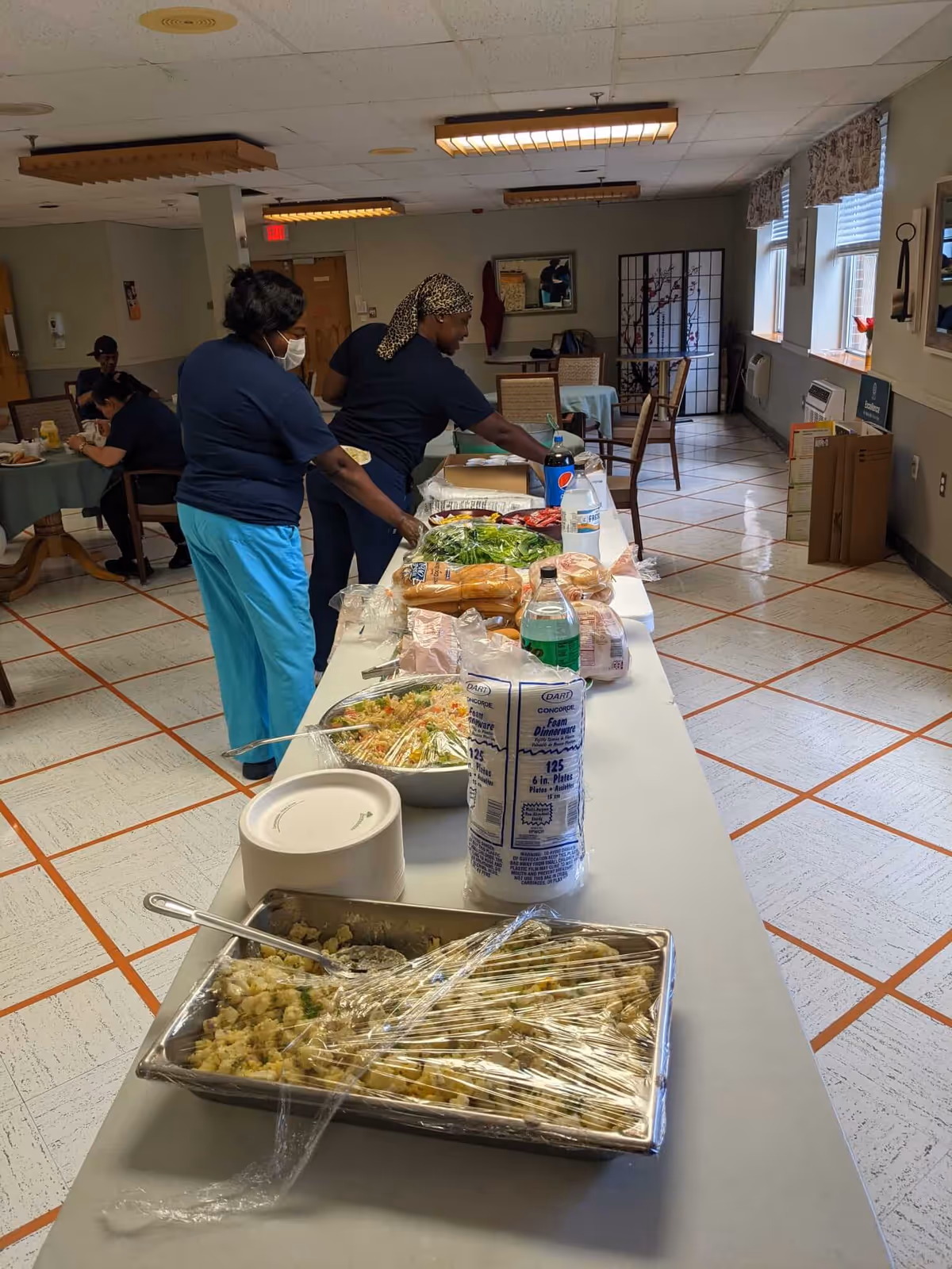 Two staff members preparing a buffet-style food table in a dining area with tables and chairs. The table has various food items including salad, bread, and drinks. Other people are seated and eating in the background.