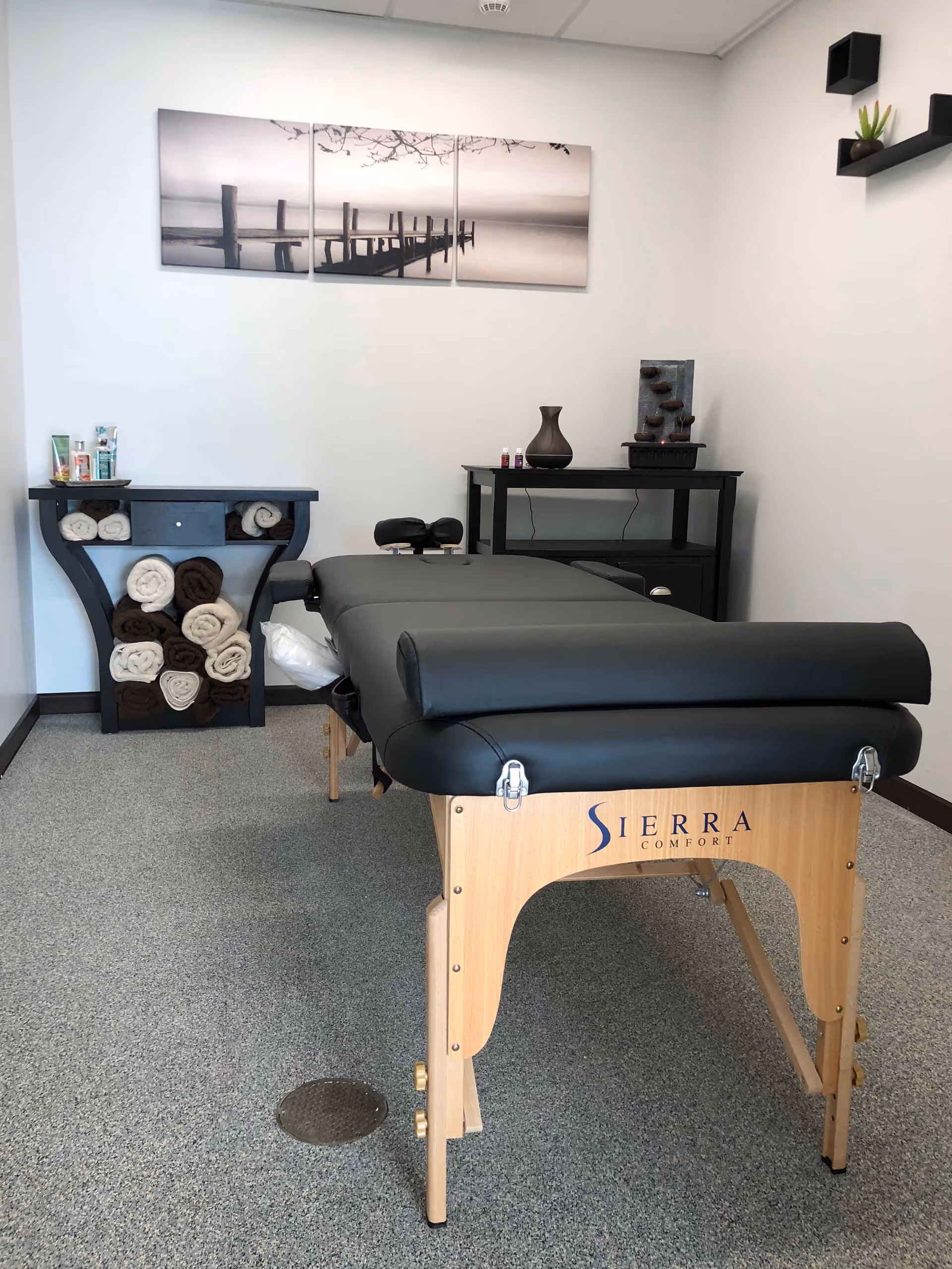 A massage therapy room with a black padded massage table labeled 'Sierra Comfort' in the center. The room has a small black table with a decorative vase and a water fountain, a shelf with rolled towels in white and brown, and a three-panel wall art depicting a pier over water in black and white. The walls are white and the floor is carpeted in gray.