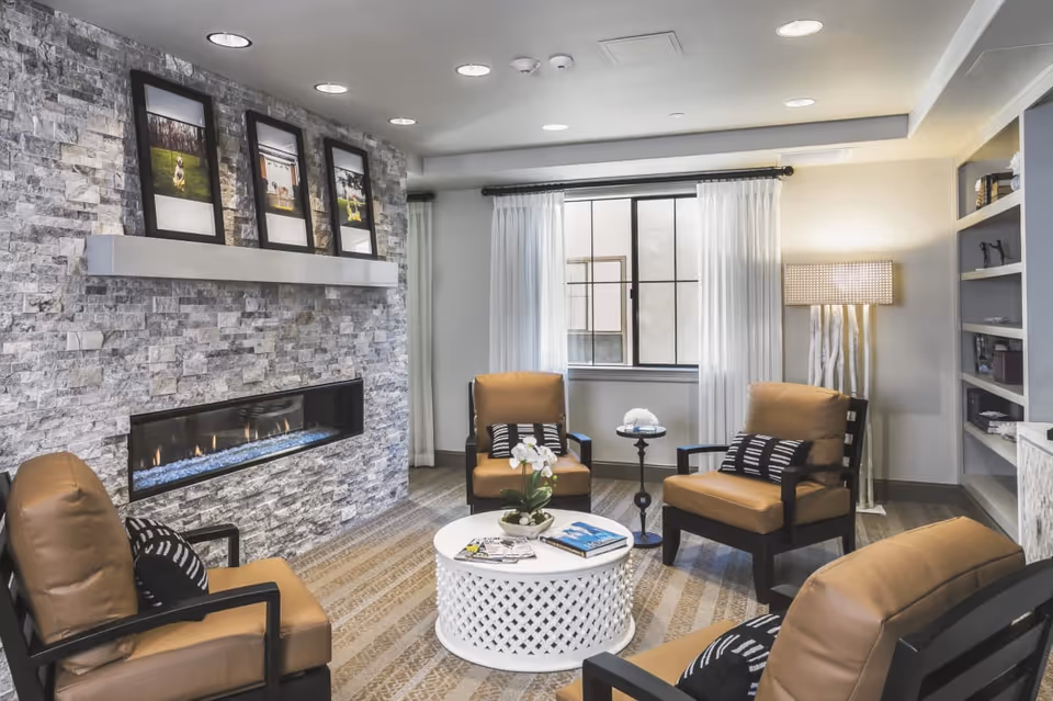 Cozy seating area with four leather armchairs around a round white coffee table, a stone accent wall with a linear fireplace and framed photos, and a window with curtains.