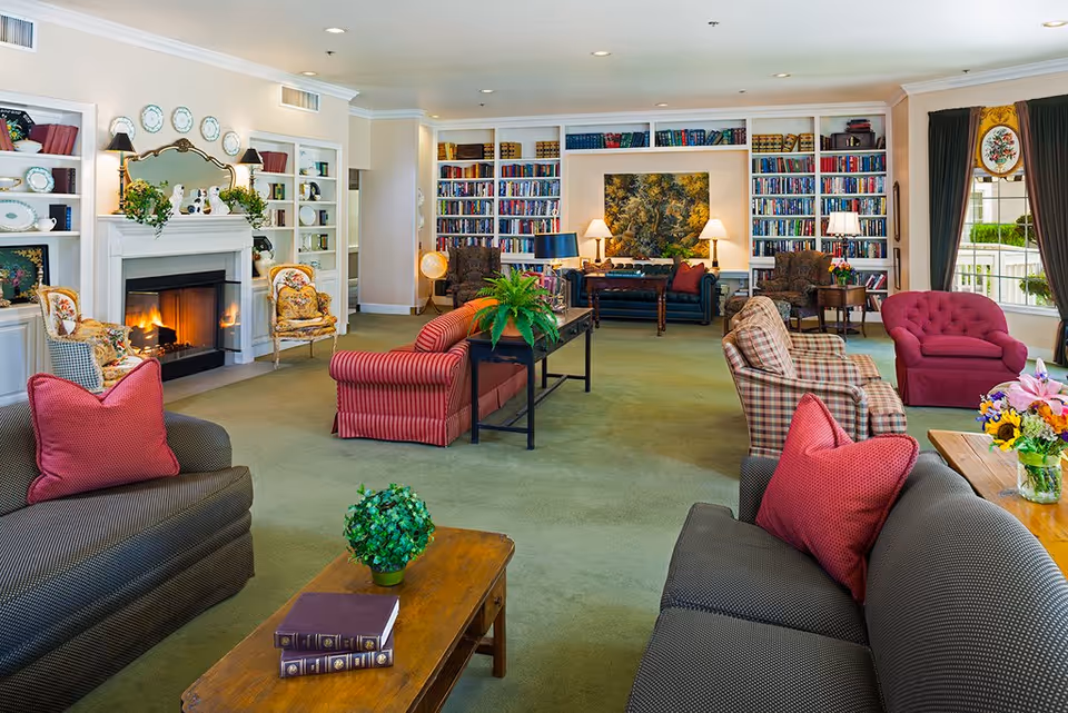 A spacious and cozy living room in an assisted living community featuring multiple upholstered chairs and sofas with red and plaid cushions, a wooden coffee table with books and a small plant, a fireplace with decorative plates and plants on the mantel, built-in bookshelves filled with books, and large windows with dark curtains allowing natural light to enter.