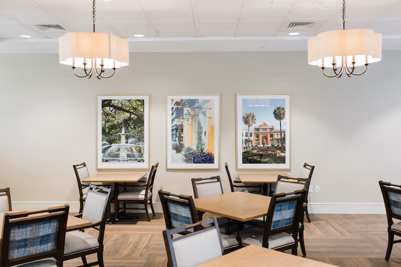 Bright dining room with wooden tables and chairs, three framed artworks on the wall, and pendant lights overhead.