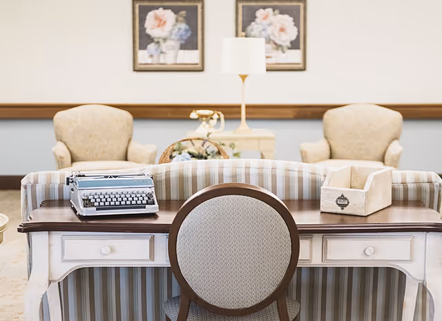 A cozy senior living room area featuring a wooden desk with a vintage typewriter and a wooden organizer on top. Behind the desk is a striped sofa, and in the background are two upholstered armchairs, a small table with a lamp, and two floral paintings on the wall.