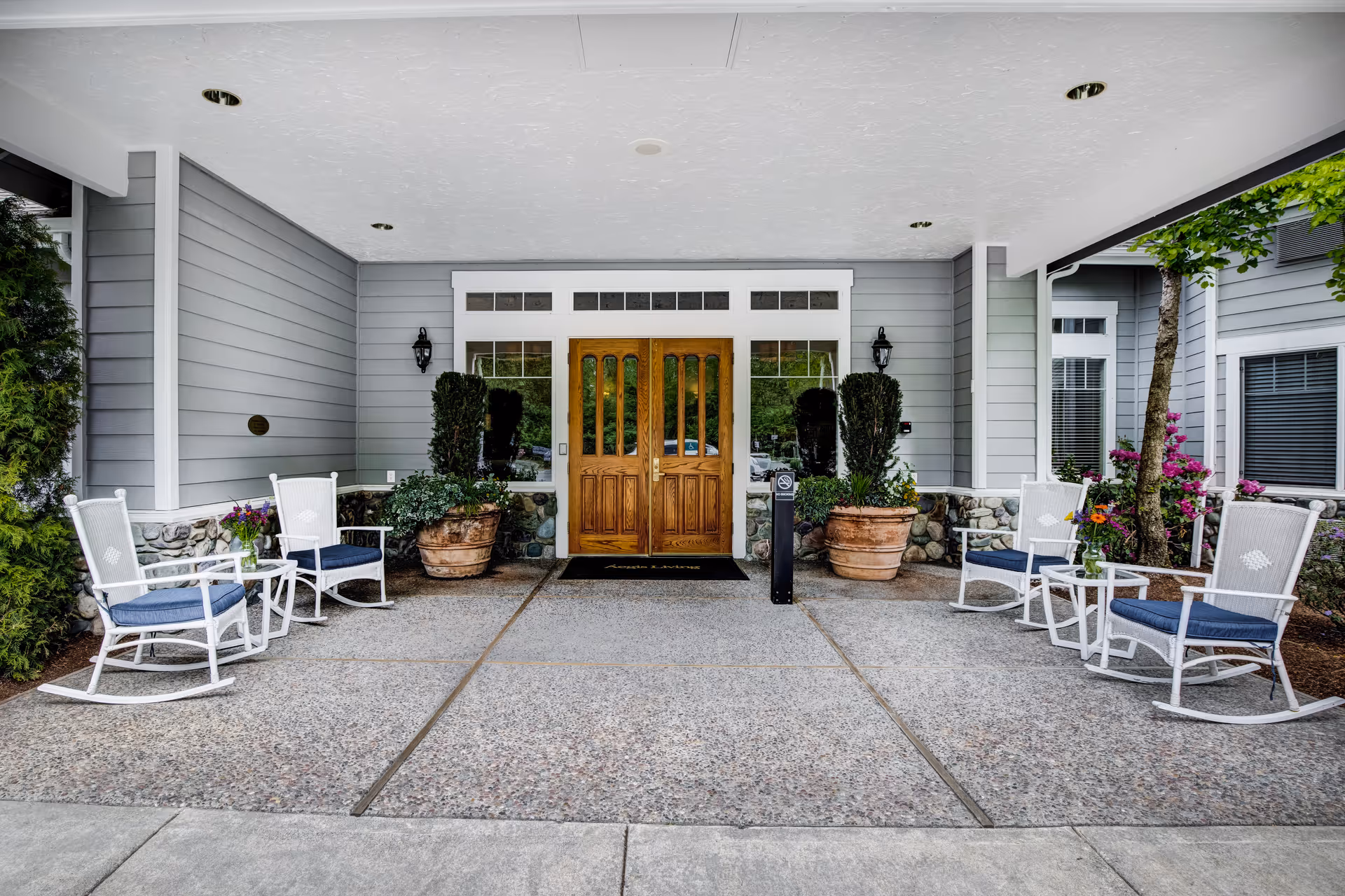 Covered entrance area of Aegis Living Callahan House featuring double wooden doors flanked by windows and two large potted plants. On either side of the entrance are white rocking chairs with blue cushions and small white tables, with some flowers placed on the tables. The exterior walls are gray with stone accents at the bottom.