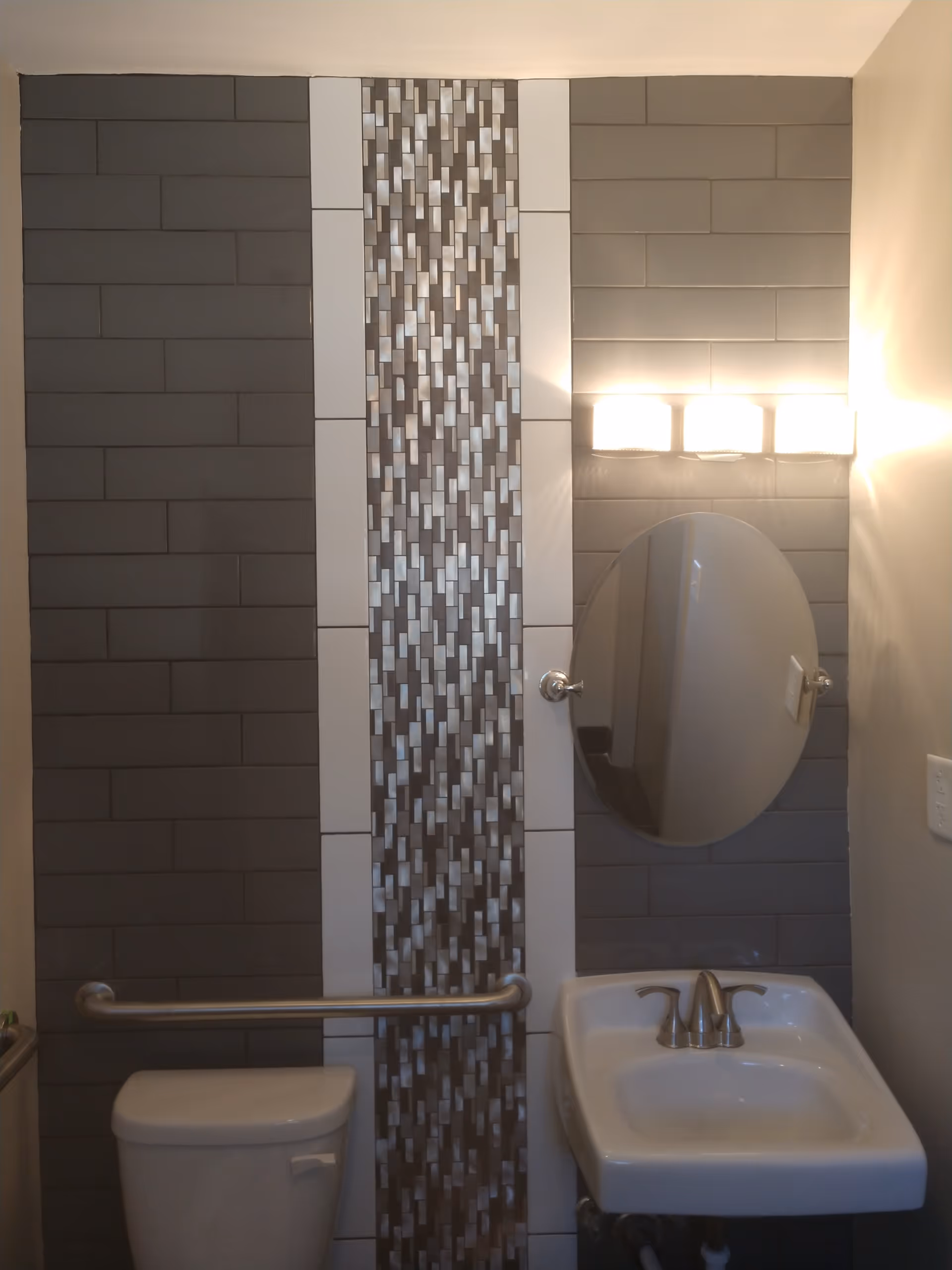 Small modern bathroom with a toilet, sink, oval mirror, wall light and a vertical mosaic tile accent within gray subway tile.