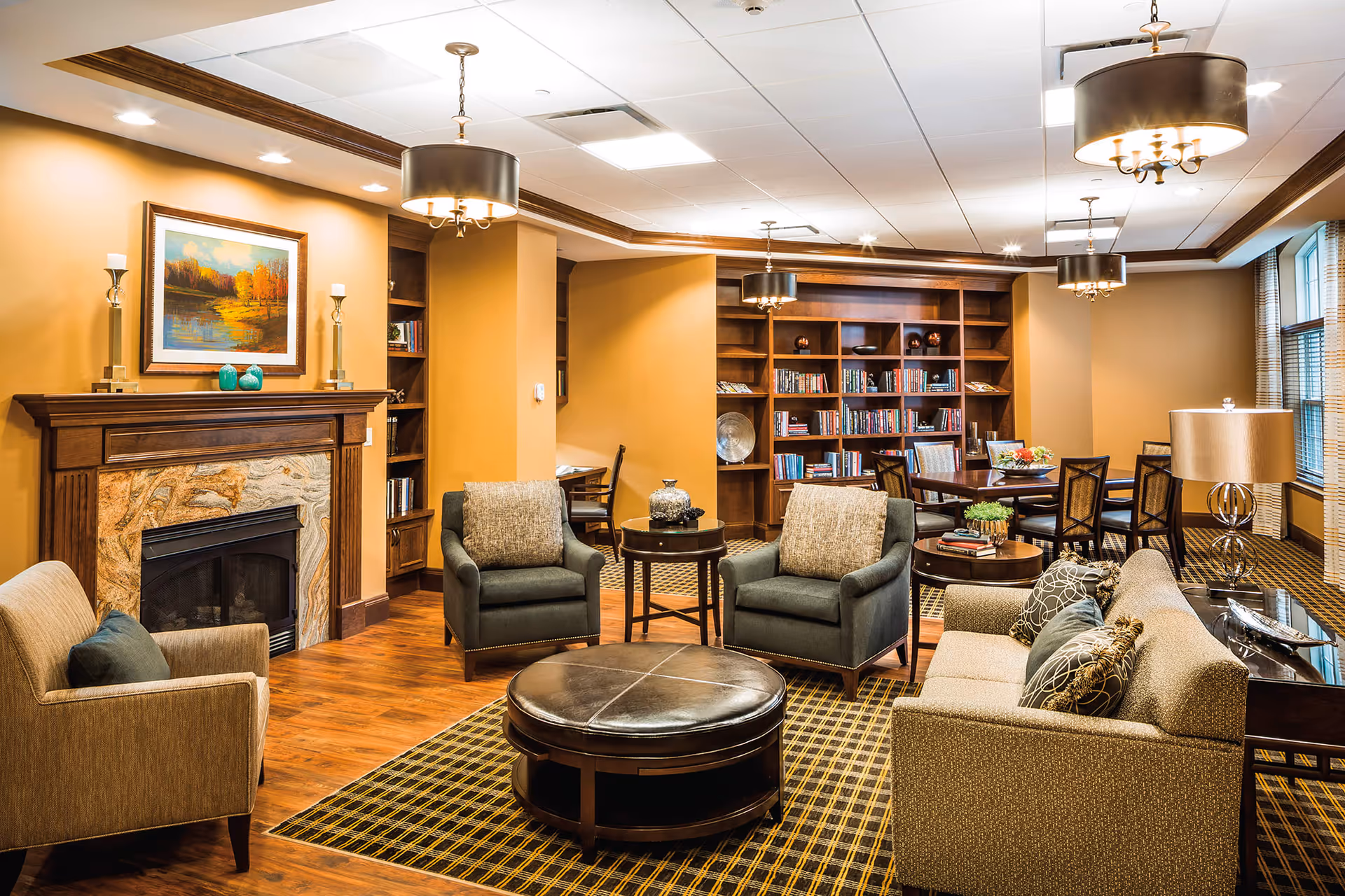 A cozy living room area in a senior living facility featuring a fireplace with a painting above it, several armchairs and a sofa arranged around a round coffee table, wooden bookshelves filled with books, a dining table with chairs, and warm lighting from ceiling fixtures and lamps.