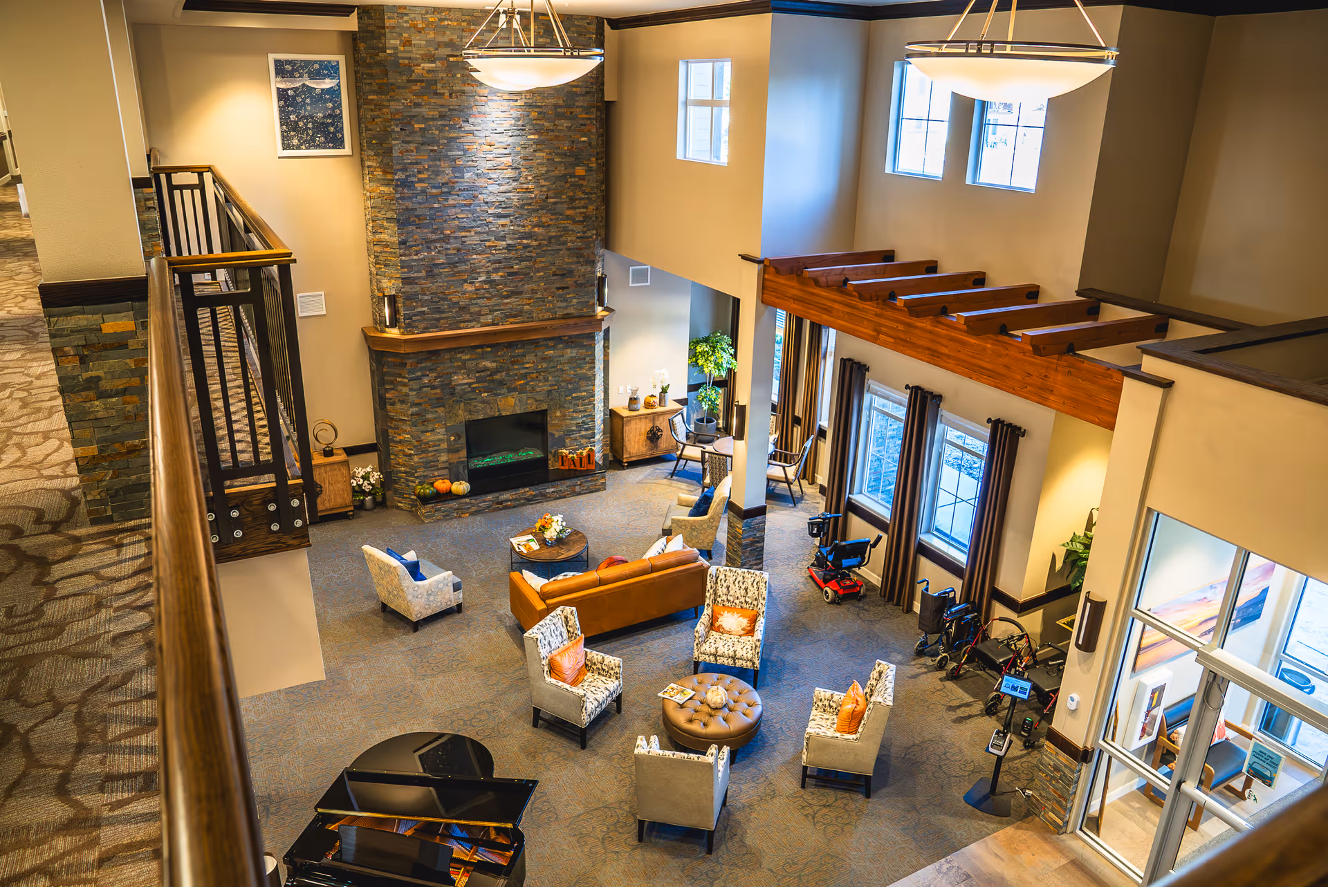 A spacious and well-lit living room area in a senior living facility with a stone fireplace, multiple armchairs, a sofa, a round ottoman, and a grand piano. Large windows with curtains allow natural light to fill the room. Mobility scooters and walkers are parked near the windows. The room features high ceilings with wooden beams and modern light fixtures.