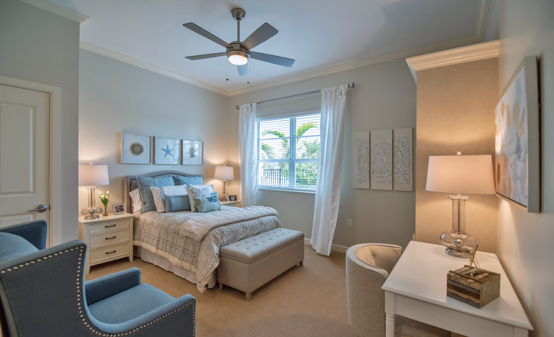 A cozy bedroom with a neatly made bed featuring multiple pillows and a patterned comforter. The room has light-colored walls, a ceiling fan, two bedside tables with lamps, a window with white curtains showing greenery outside, a blue armchair, a white desk with a lamp and decorative items, and wall art including framed pictures and carved panels.